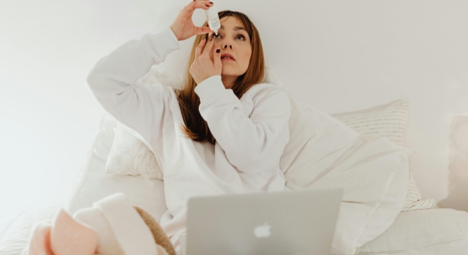 Person in white hoodie applying eye drops while sitting on a bed with a laptop.