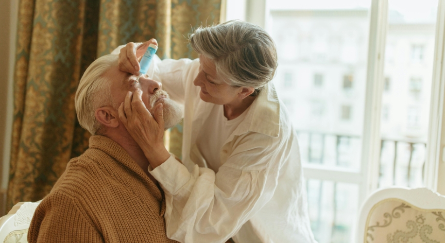 Woman applying eye drops to an older man seated in a chair.