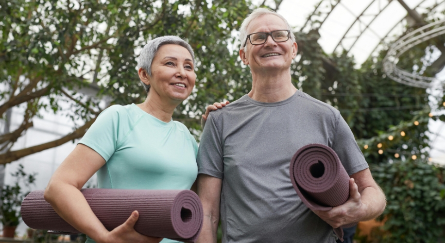 Two people holding rolled yoga mats with smiles on their faces, standing in a garden.