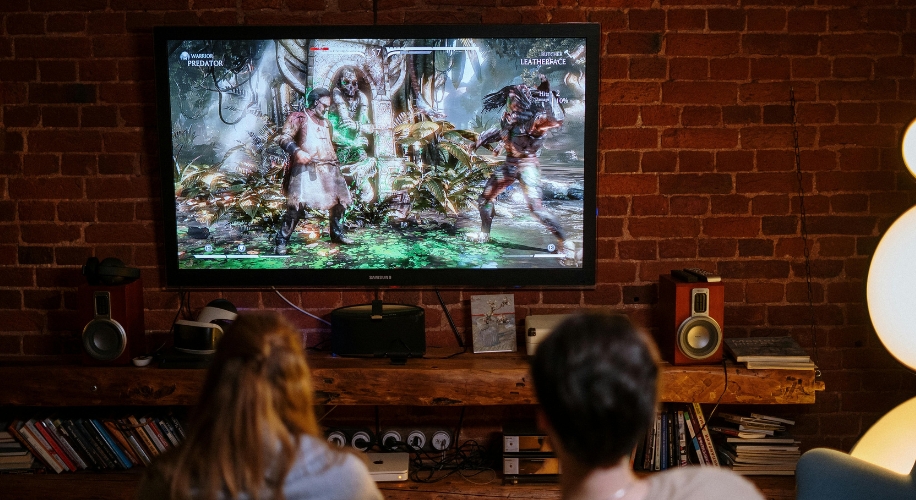 People playing a fighting video game on a large TV screen. Characters: Marauder Predator and Leatherface.