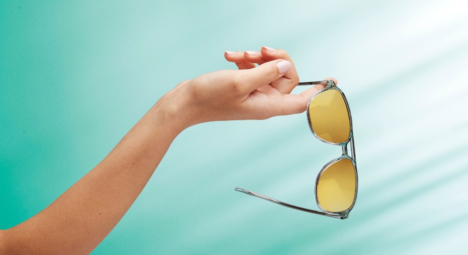 Hand holding yellow-tinted aviator sunglasses with a silver frame against a turquoise background.