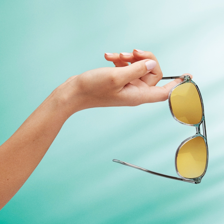 Hand holding sunglasses with yellow-tinted lenses and a clear frame against a teal background.