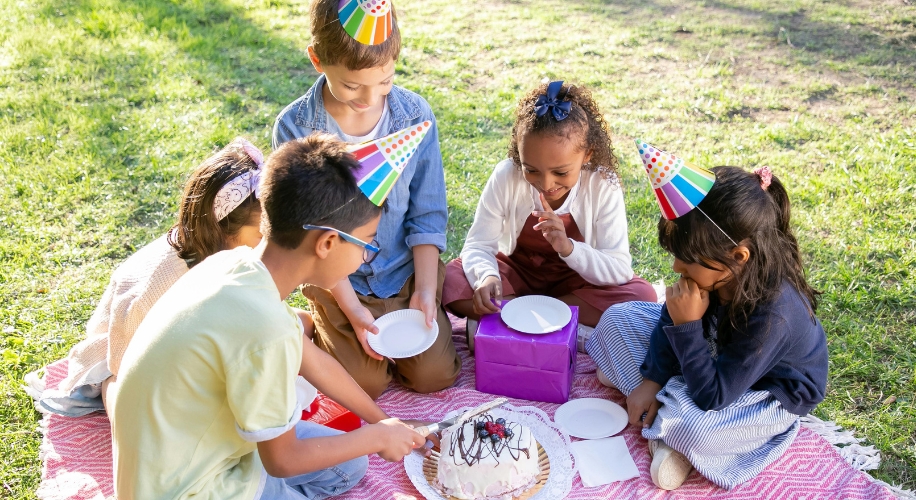 Children wearing party hats gathered around a cake on a picnic blanket, holding plates and smiling.