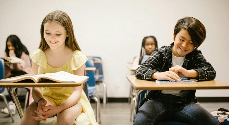 Two smiling kids sitting at desks in a classroom, reading books.