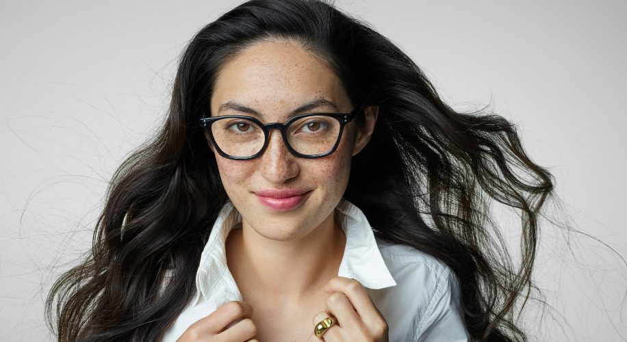 Woman wearing black-framed glasses and a white shirt. She has long, wavy dark hair.