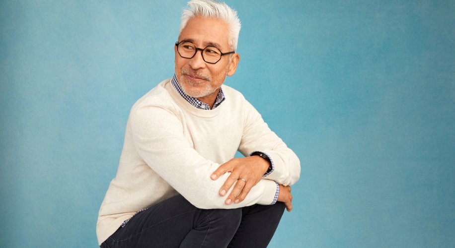 Man wearing a light-colored sweater, patterned shirt, and dark jeans squatting against a blue background.