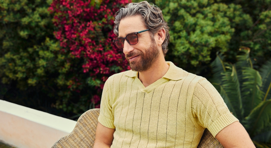 Man wearing a light yellow knit polo shirt and brown sunglasses, sitting outdoors.