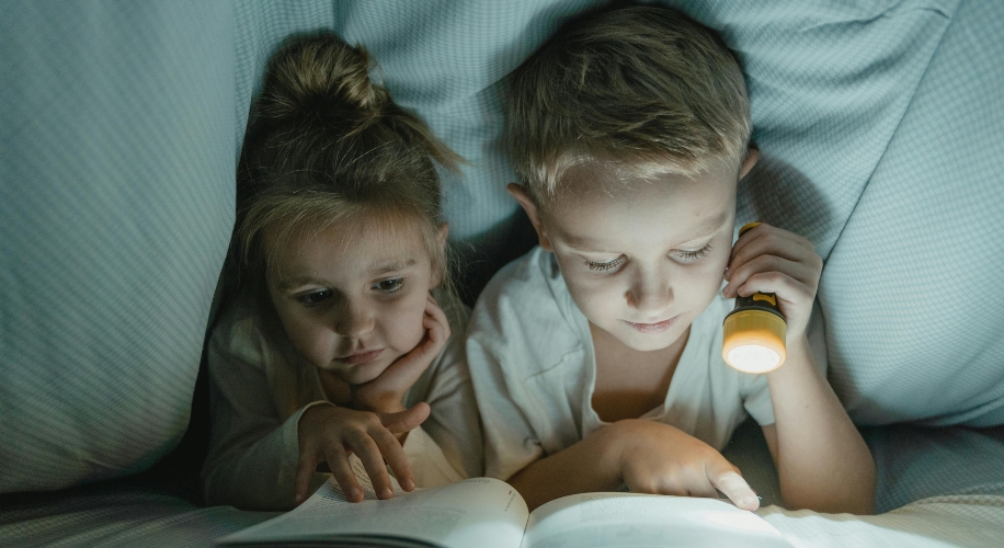 Two children reading a book under a blanket, illuminated by a yellow flashlight.