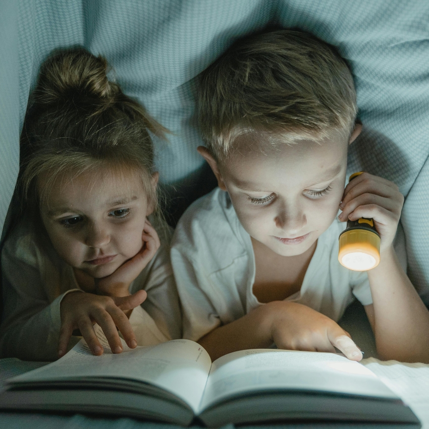 Children reading a book under a blanket with a yellow flashlight illuminating the pages.