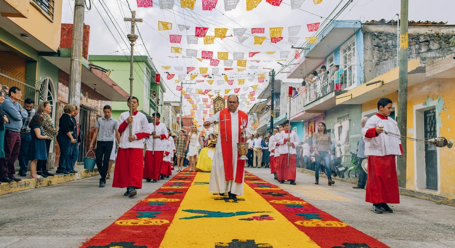 Religious procession with priests and altar servers wearing red and white vestments on a colorful street carpet.