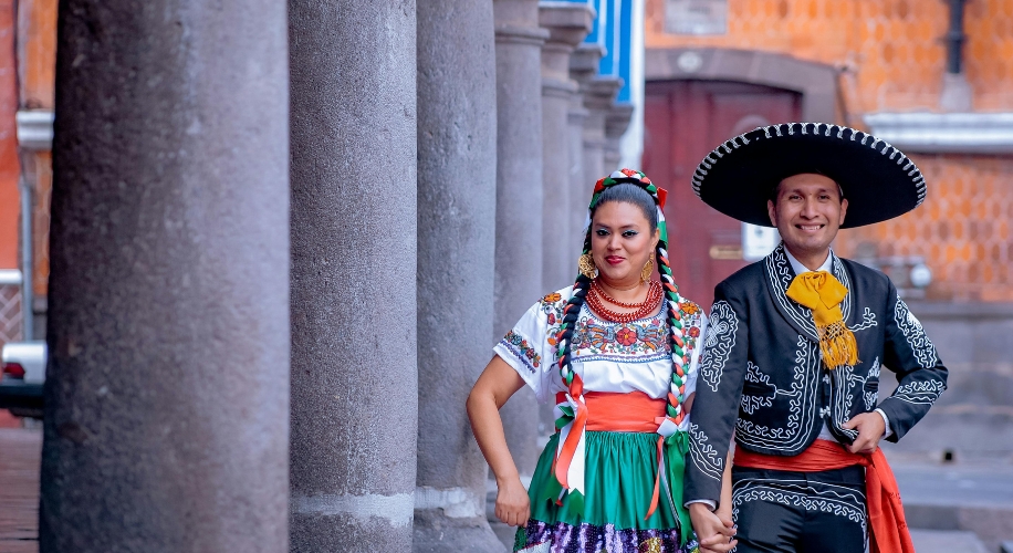 Two people in traditional Mexican folk dance attire, standing between stone columns.