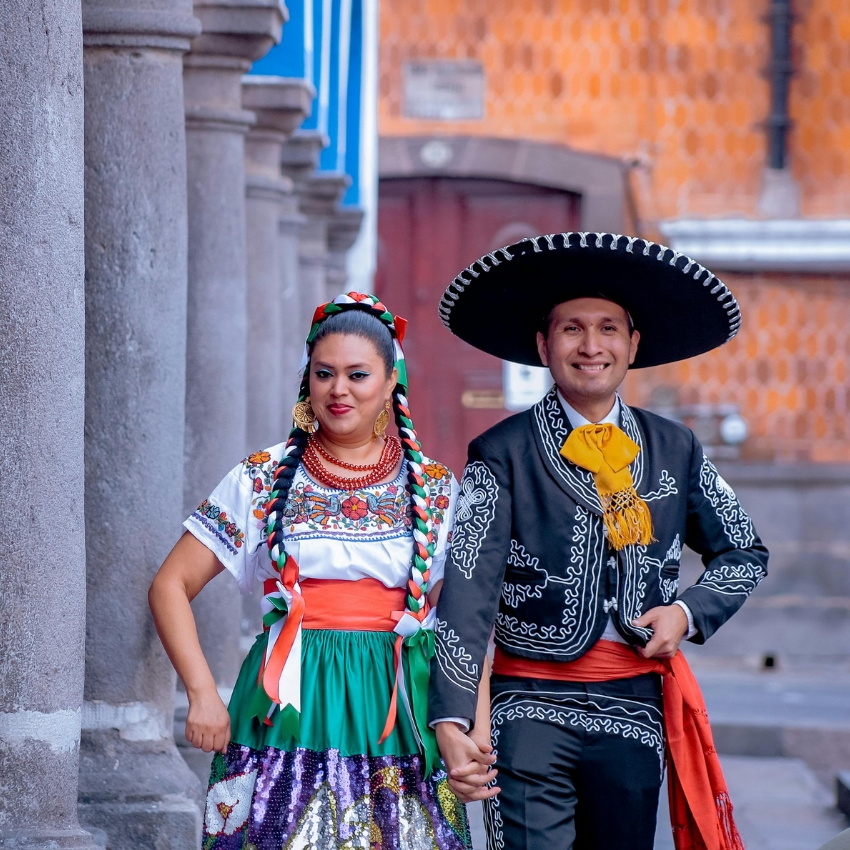 Couple wearing traditional Mexican clothing; woman in colorful embroidered dress and braided hair, man in black charro suit with sombrero.