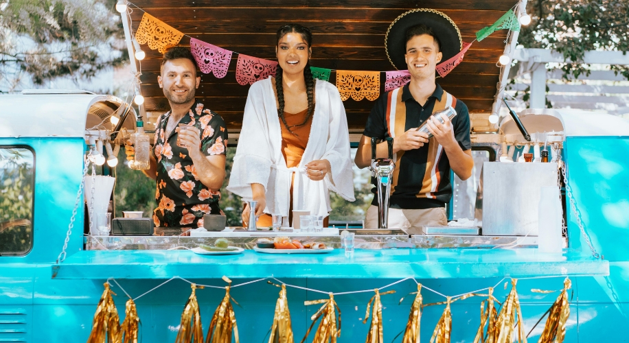 Three people at a festive food truck counter, two holding drinks and one holding a cocktail shaker.