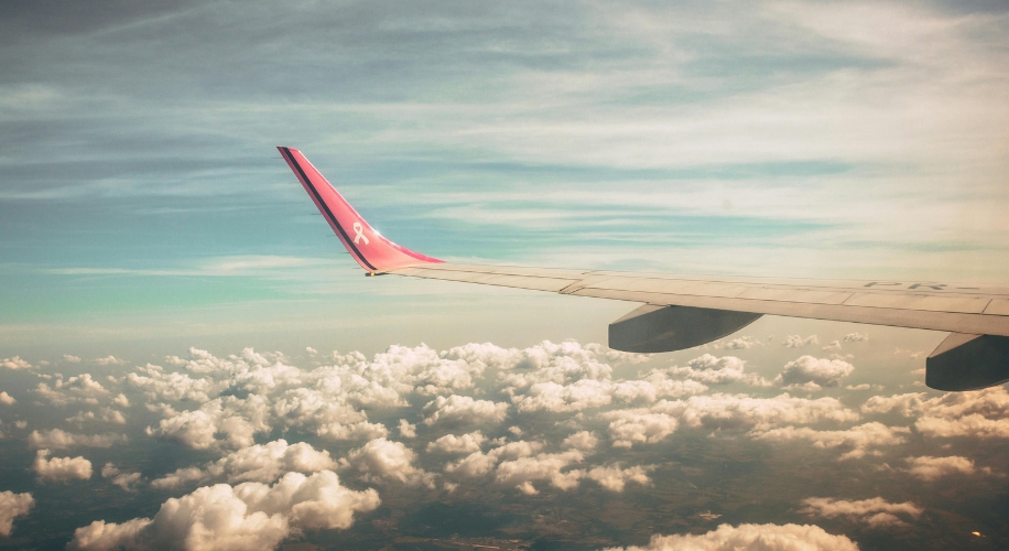 Airplane wing with a pink ribbon on the winglet, flying above clouds under a blue sky.