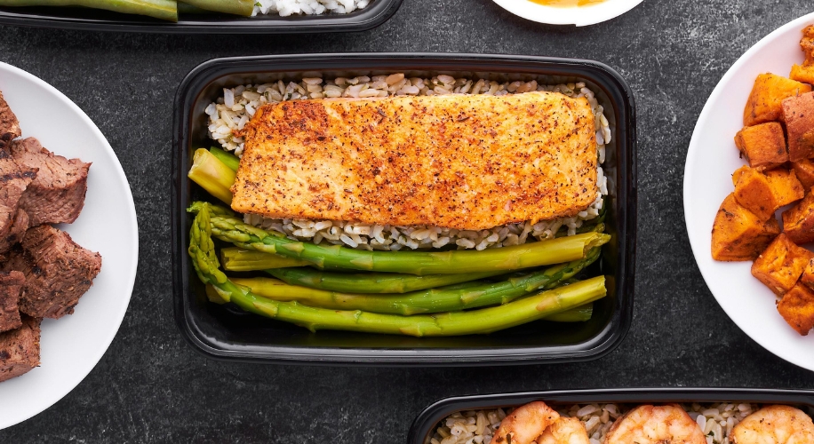 Seasoned salmon fillet on a bed of rice with asparagus in a black container.