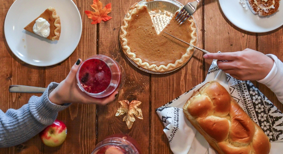 Pumpkin pie with a slice removed, a slice on a plate with whipped cream, braided bread, and a cranberry drink.