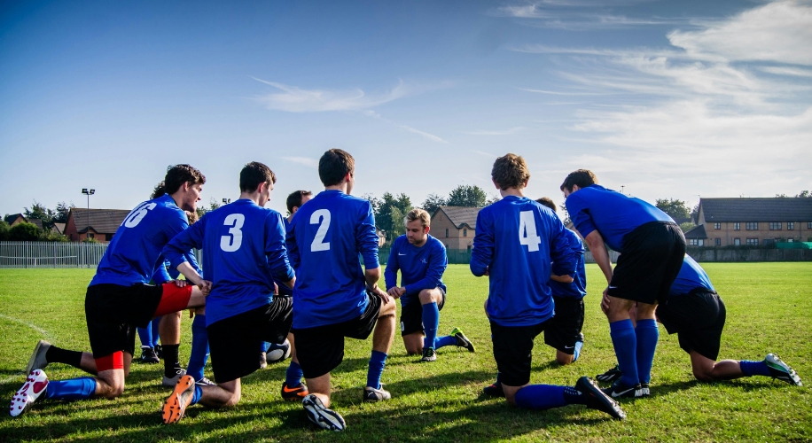 Soccer team in blue jerseys with numbers kneeling on a field while discussing strategy.