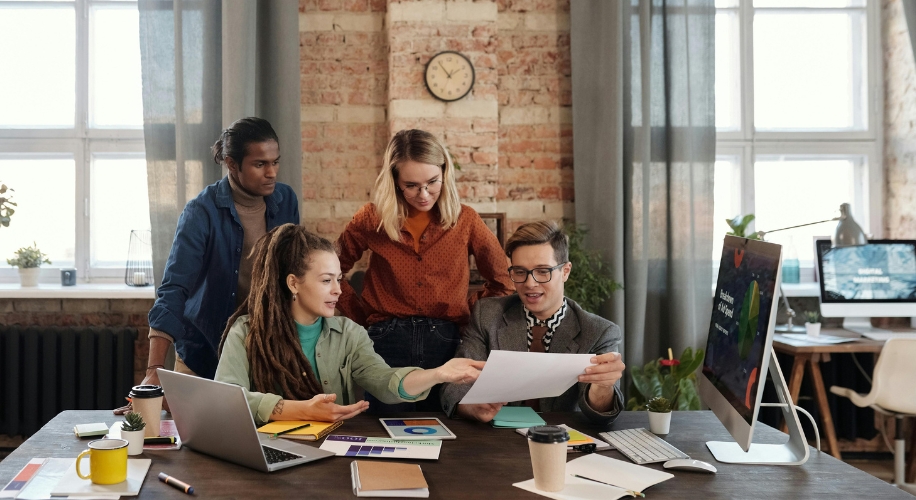 Four people collaborating at a desk with documents, a laptop, a smartphone, a tablet, and a monitor.