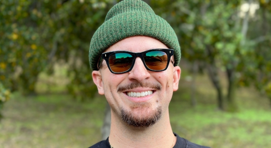 Man wearing dark sunglasses and a green knit beanie, smiling outdoors.