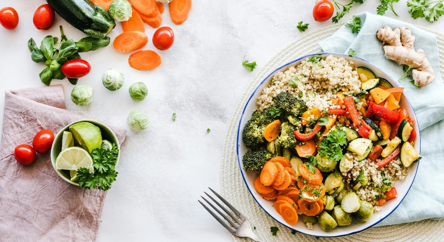 Bowl of quinoa, broccoli, carrots, zucchinis, and bell peppers. Sliced vegetables and lime wedges nearby.