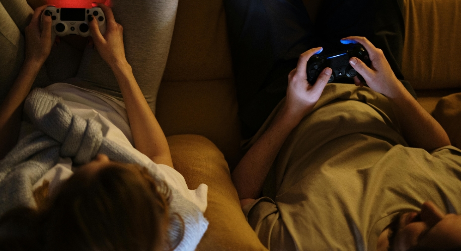 Two people holding and playing video game controllers while lying on a couch.