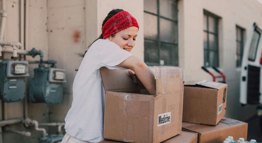 Woman in white shirt and red bandana putting items in a cardboard box labeled &quot;Medicine.&quot;