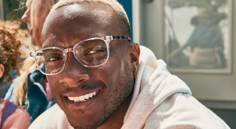 Man wearing clear-framed glasses and a white hoodie, smiling outdoors.