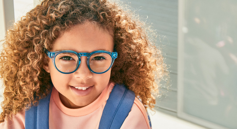 Child wearing blue-framed eyeglasses and light pink shirt with a blue strap from a backpack.