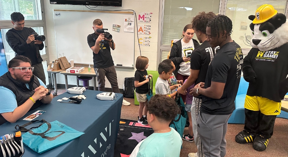 Man at a table with 4D Replay branding, surrounded by people, two cameras, and a costumed mascot.