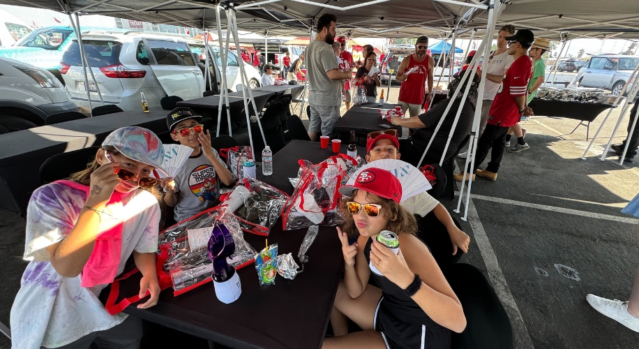 People enjoying snacks and drinks under canopies, wearing San Francisco 49ers apparel and sunglasses.