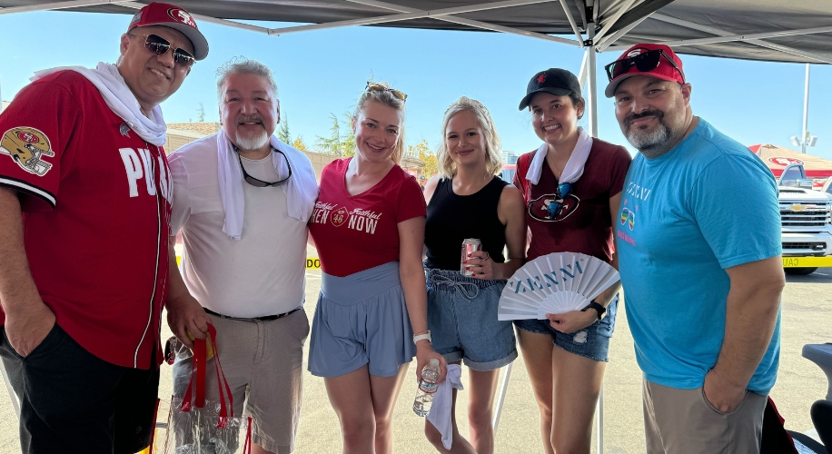 Six people standing together; two wear red 49ers shirts, one holding a fan and another a bottle.