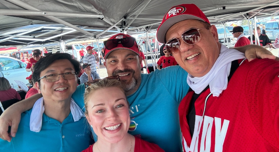 Four people smiling under a tent. Shirt text partially visible: &quot;JOY.&quot;