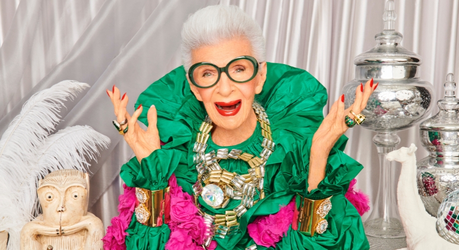 Woman in a vibrant green and pink outfit with bold jewelry and large round glasses.