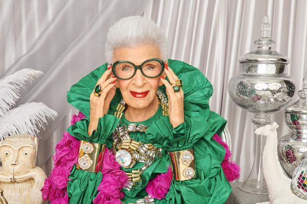 Woman wearing oversized green dress with pink flowers, large glasses, and bold jewelry.