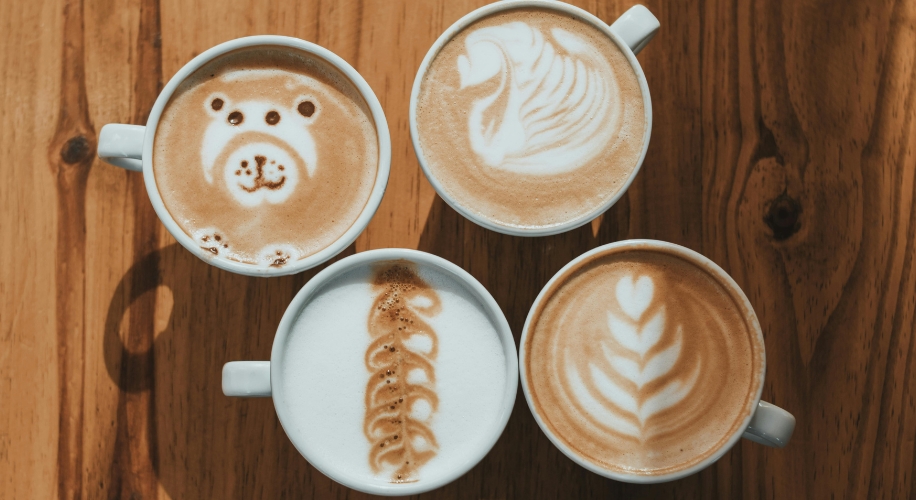 Four cups of coffee with latte art: a bear, a swan, a fern, and a heart design on frothy milk.