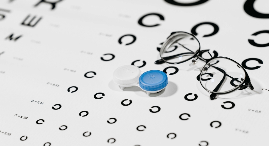 Contact lens case with blue and white caps beside eyeglasses on an eye test chart.