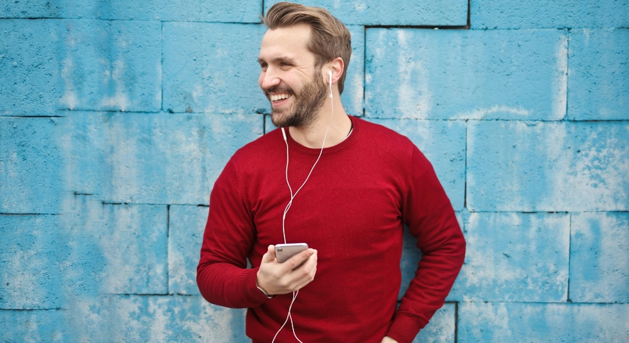 Man wearing a red sweater using white earphones with a smartphone against a blue brick wall.