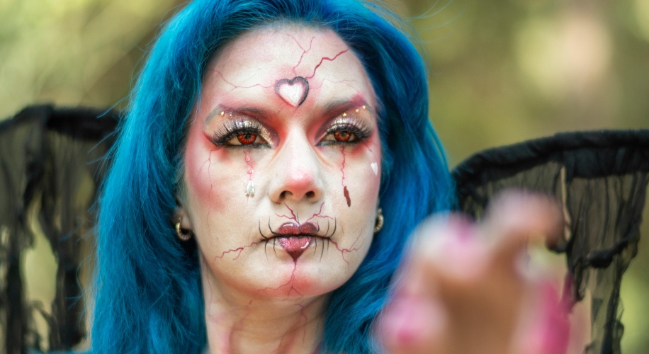 Fantasy makeup with blue hair, heart design on forehead, red and black accents, and dramatic wings.