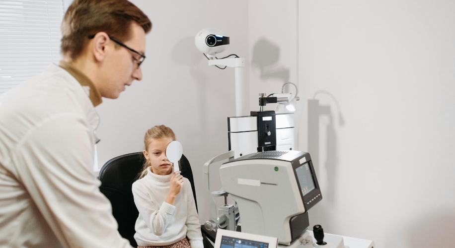 Child undergoing an eye examination with an autorefractor machine, supervised by an optometrist.