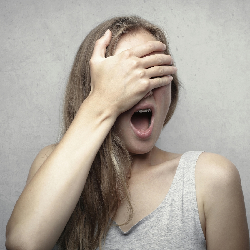 Woman with long hair covering her eyes with one hand, mouth open.