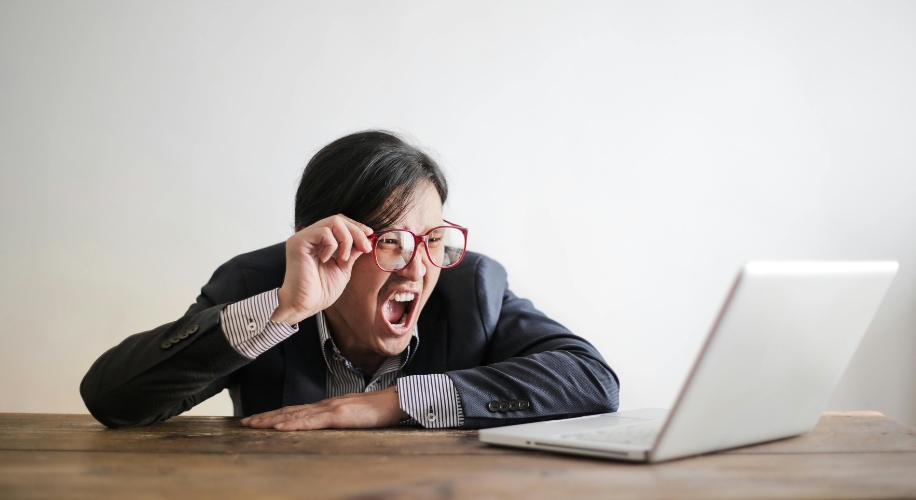 Person wearing glasses, shouting at a laptop on a wooden table.