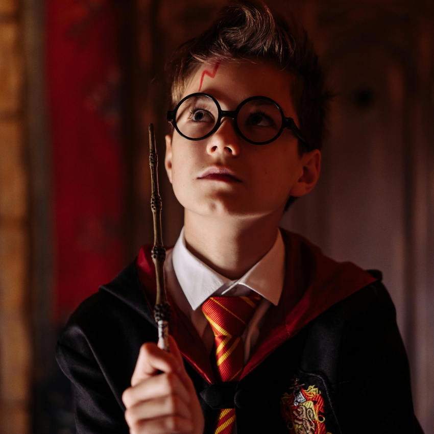 Young person wearing round glasses, holding a wand, and dressed in a robe with a striped tie.