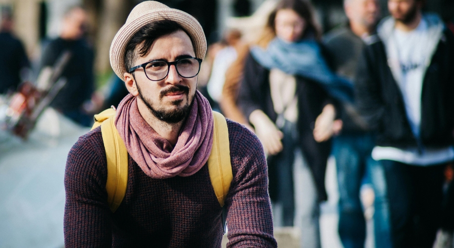 Man wearing glasses, hat, pink scarf, yellow backpack, and a dark sweater.