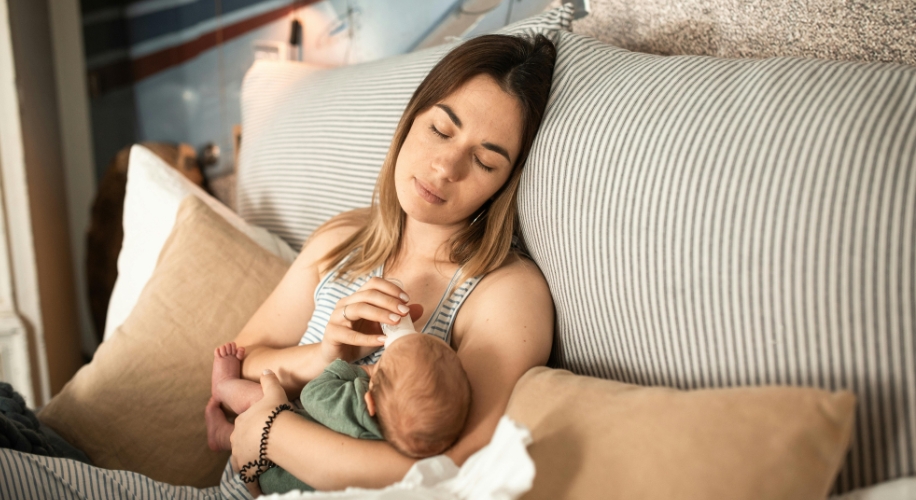 Woman nursing a baby on a striped bed with neutral-colored pillows.