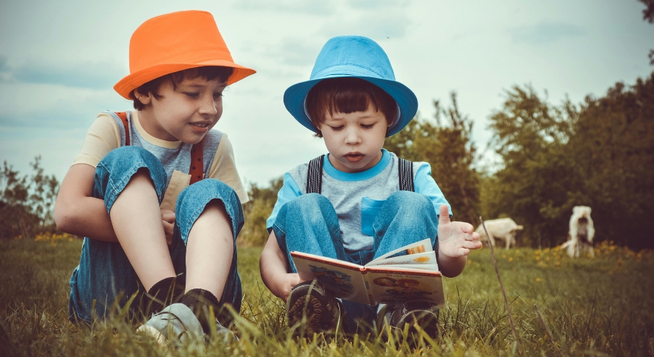 Two children in colorful hats reading a book outdoors.