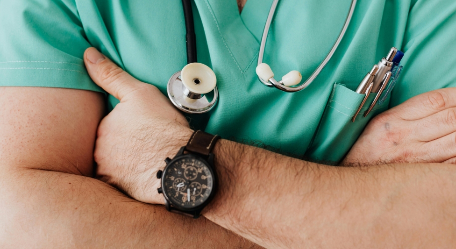 Stethoscope on torso in green medical scrubs, arm folded with a black watch and pens in pocket.