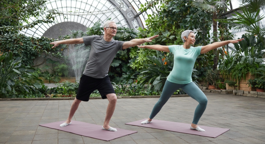 Two people practicing yoga on mats, holding a warrior pose inside a greenhouse.