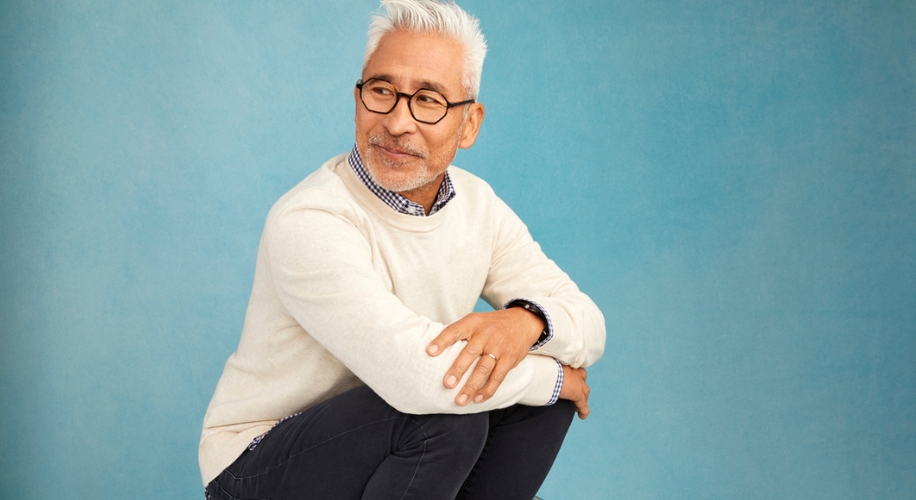 Man wearing glasses, cream sweater, and dark pants against a blue background.