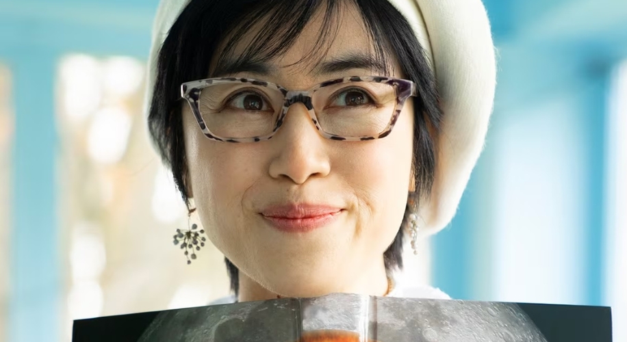 Woman wearing patterned glasses and dangle earrings, smiling with a book in front.