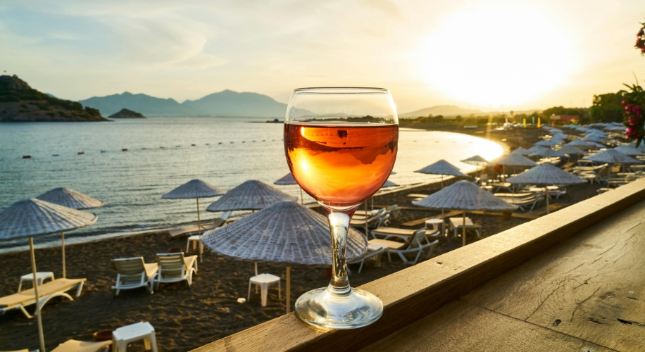 Glass of rosé wine on a wooden railing by the beach at sunset.